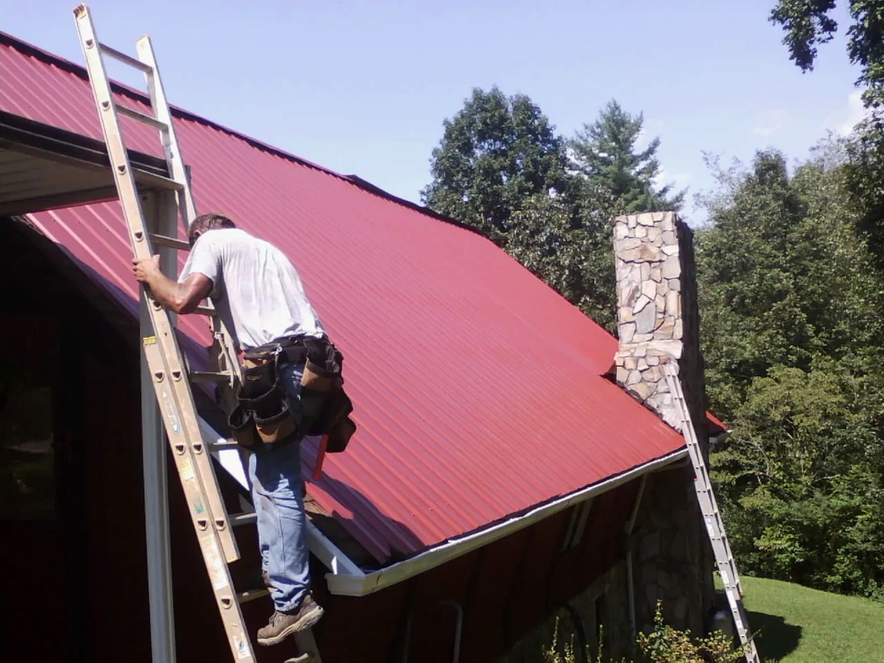 Roofer on a ladder beside a red metal roof and stone chimney — Soffit & Fascia Repair by Roofing Brothers in Lake Arrowhead